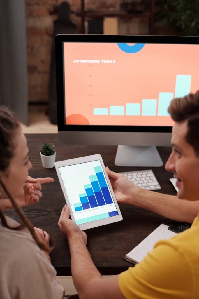 A team of colleagues discussing sales growth using a digital tablet and computer screen in an office setting.